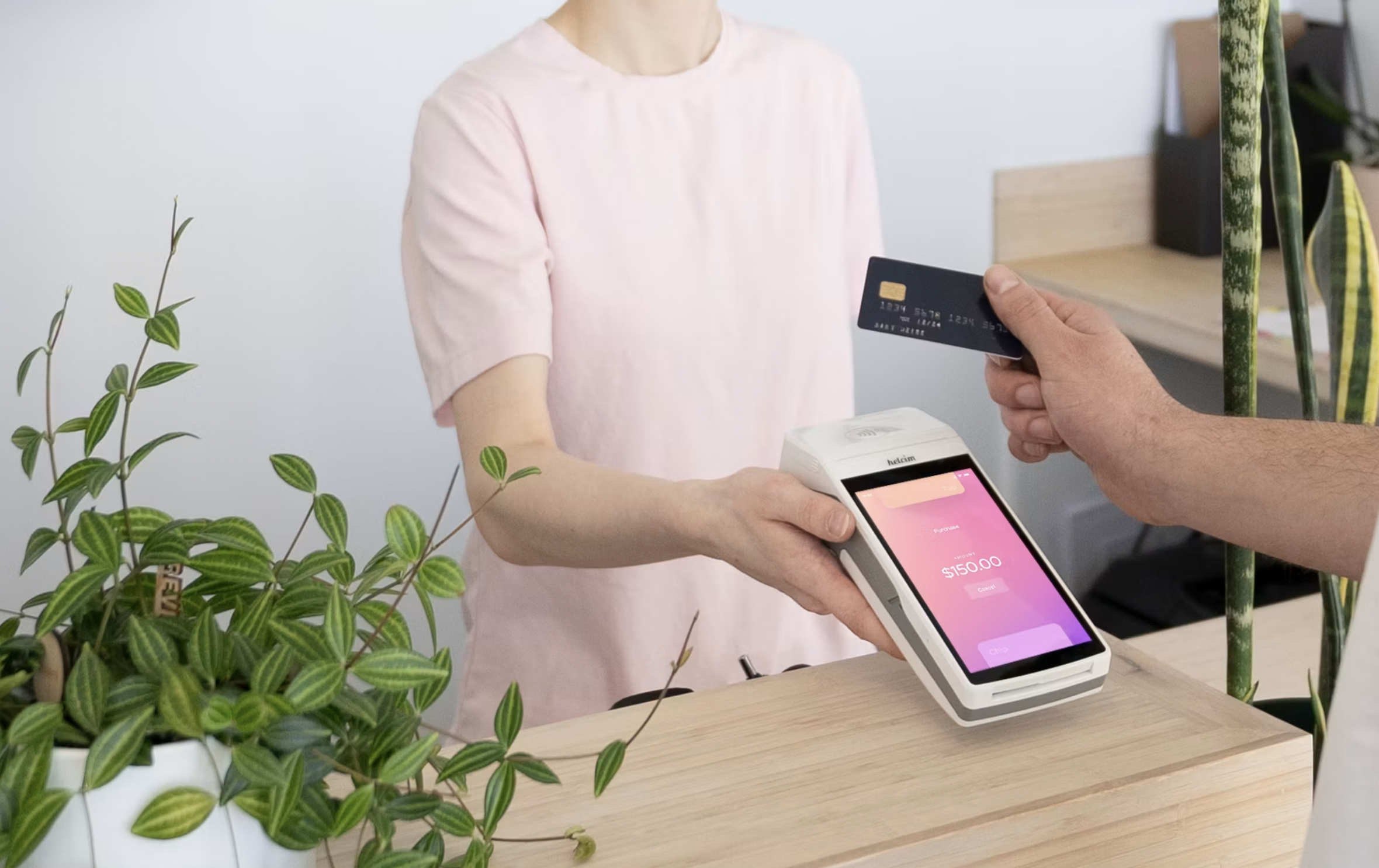 Customer hands a card to the cashier at a checkout, with the card reader showing $150.00 on screen.