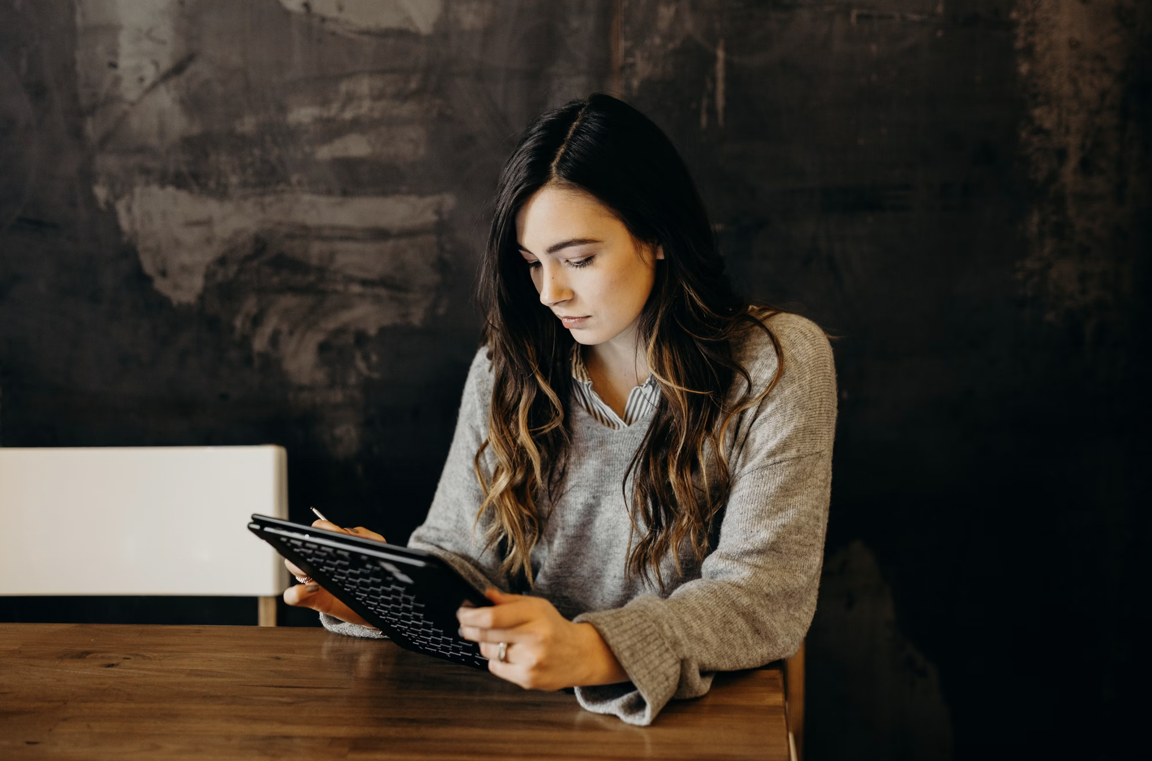 Woman in a gray knit sweater sits at a wooden table, looking down at a keyboard-like device she’s holding.
