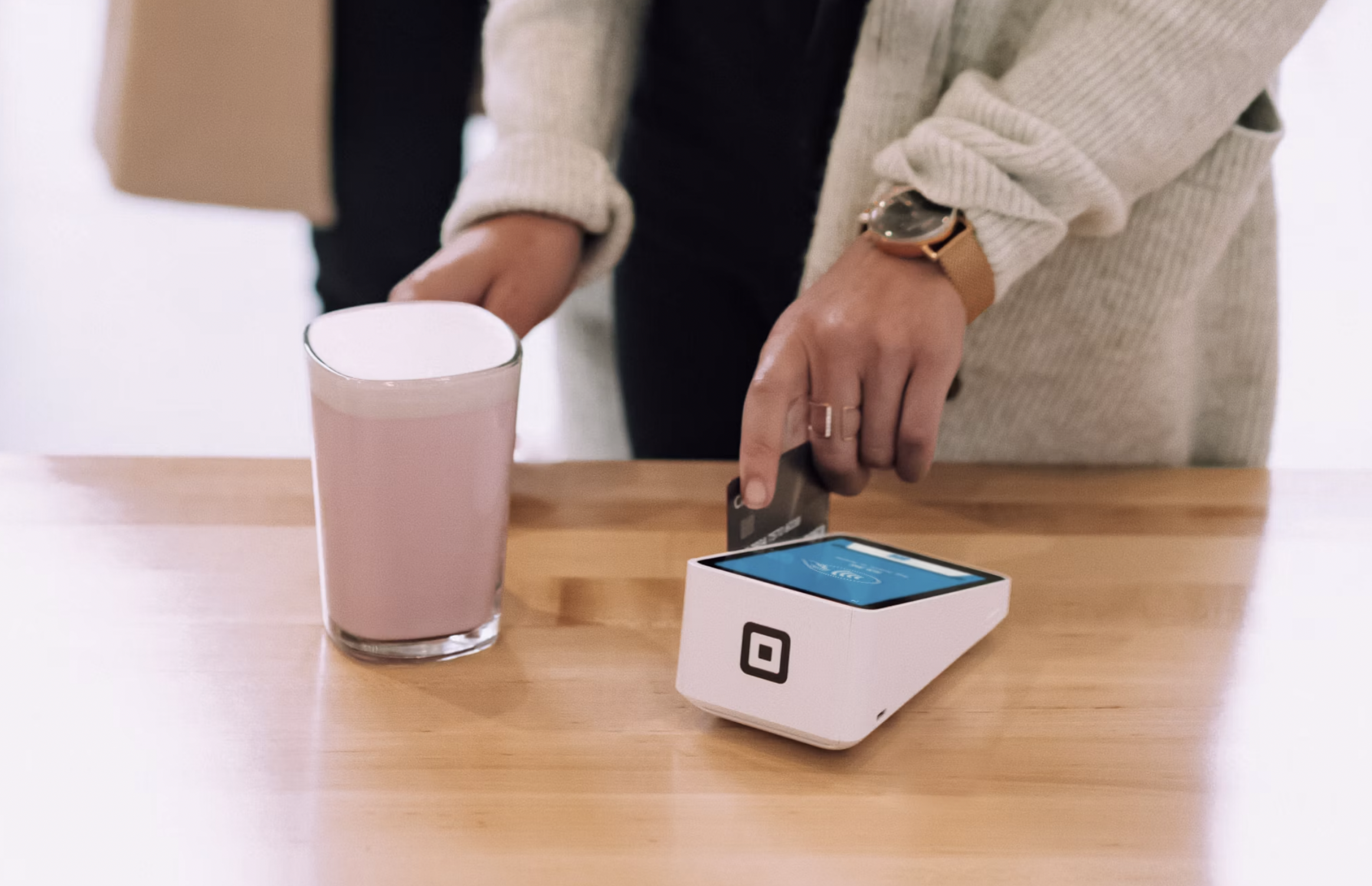 Person wearing a beige sweater and gold watch taps a card on a small white payment terminal on a wooden table, with a pink cup nearby.