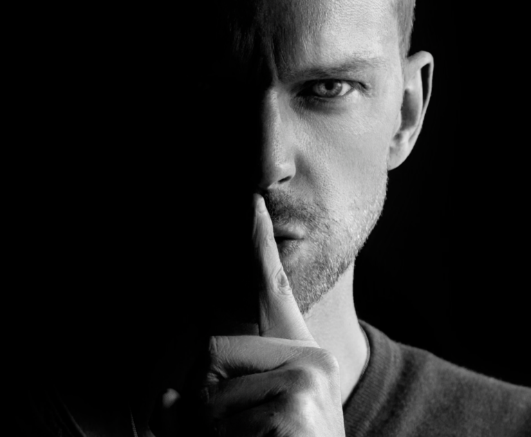 Close-up of a man's face in black and white, with his finger pressed to his lips in a shushing gesture.