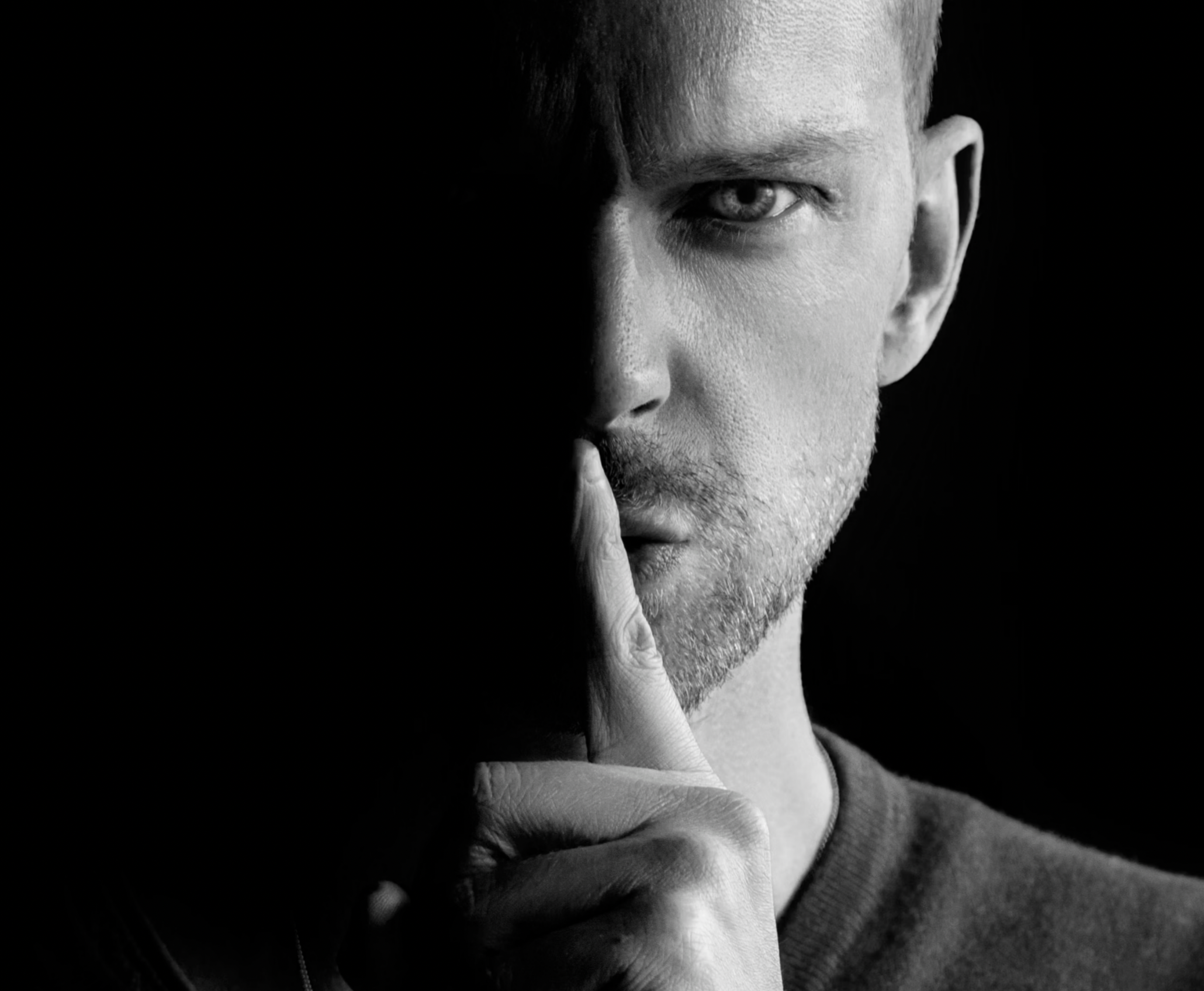 Close-up of a man's face in black and white, with his finger pressed to his lips in a shushing gesture.
