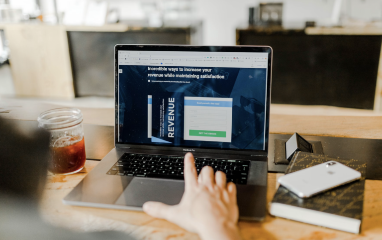 Person using a laptop at a wooden desk with a glass of iced tea nearby, screen shows a marketing landing page about increasing revenue.