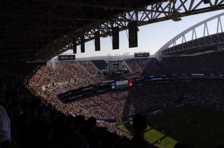 A packed soccer stadium filled with spectators under a large steel roof, with banners reading SEATTLE and FIFA World Cup 2026 along the stands.