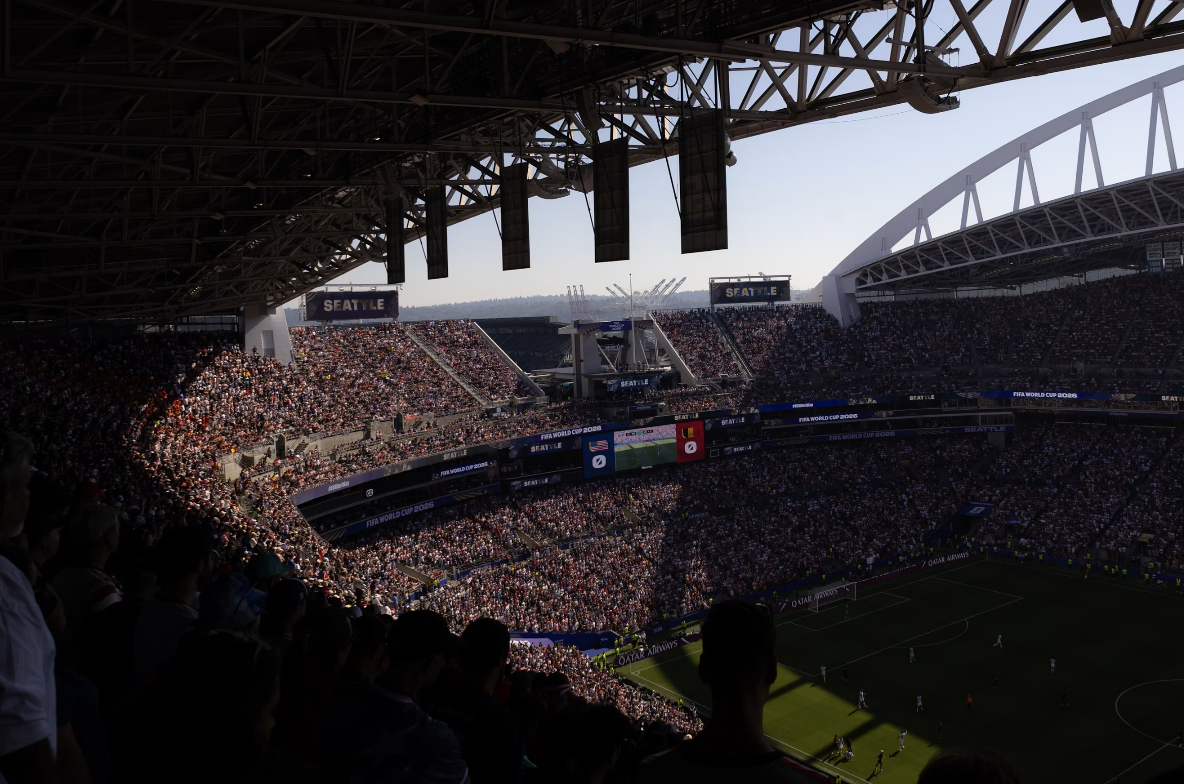 A packed soccer stadium filled with spectators under a large steel roof, with banners reading SEATTLE and FIFA World Cup 2026 along the stands.